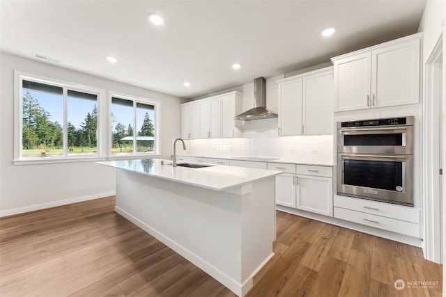 kitchen featuring double oven, sink, white cabinets, a kitchen island with sink, and wall chimney range hood