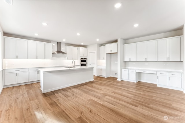 kitchen with white cabinetry, built in desk, light hardwood / wood-style flooring, and wall chimney range hood