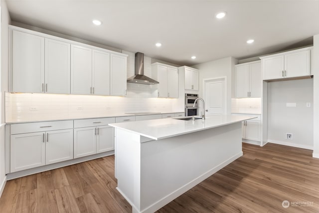 kitchen with white cabinetry, wall chimney range hood, wood-type flooring, and an island with sink