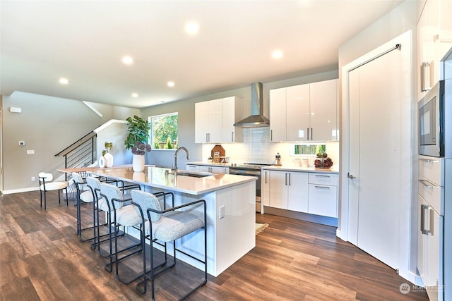 kitchen with wall chimney range hood, sink, stainless steel range with electric stovetop, a kitchen island with sink, and white cabinets