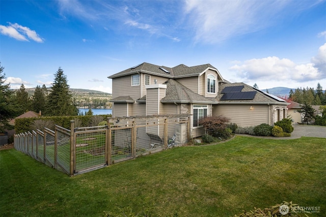 view of property exterior with a water view, fence, roof with shingles, roof mounted solar panels, and a yard