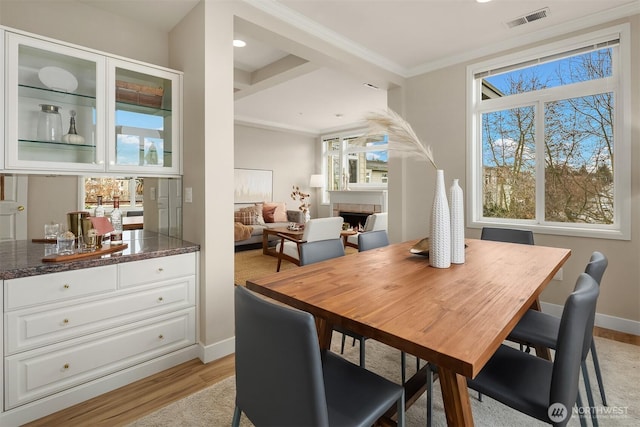 dining area with light wood-type flooring, visible vents, ornamental molding, a lit fireplace, and baseboards