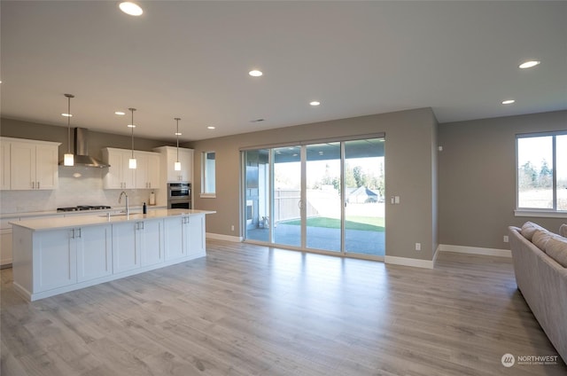 kitchen with a center island with sink, wall chimney exhaust hood, hanging light fixtures, and white cabinets