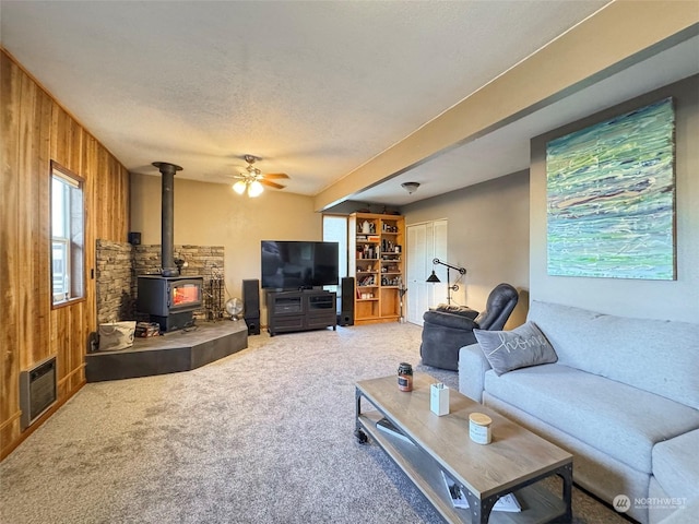 living room featuring carpet, a wood stove, wooden walls, ceiling fan, and a textured ceiling