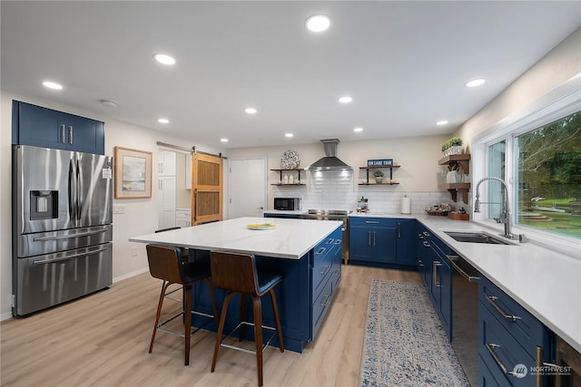 kitchen featuring sink, wall chimney exhaust hood, blue cabinetry, a barn door, and appliances with stainless steel finishes