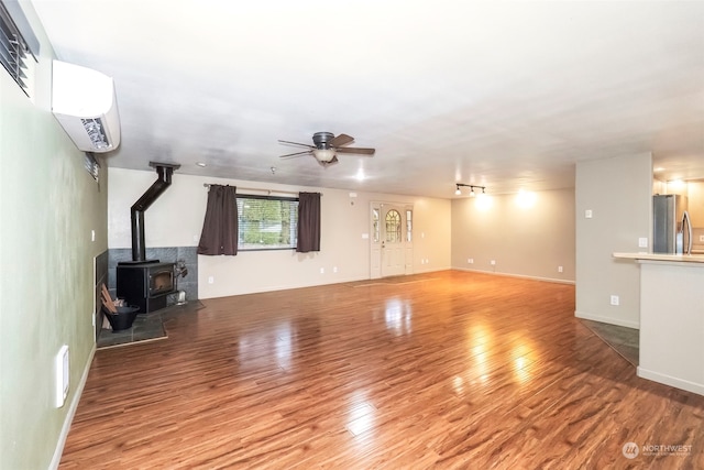 unfurnished living room featuring a ceiling fan, a wood stove, an AC wall unit, wood finished floors, and baseboards