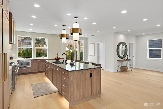 kitchen featuring a kitchen island with sink, hanging light fixtures, light wood-type flooring, dark stone counters, and sink