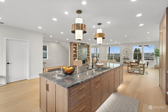 kitchen with hanging light fixtures, an island with sink, light hardwood / wood-style floors, dark stone countertops, and sink