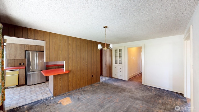 interior space with an inviting chandelier, freestanding refrigerator, brown cabinets, wooden walls, and pendant lighting