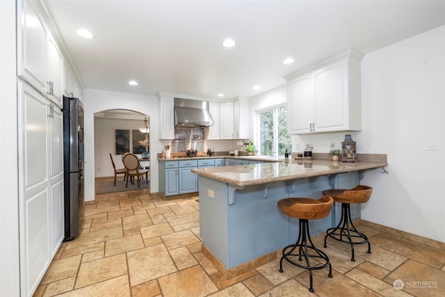 kitchen with kitchen peninsula, refrigerator, wall chimney exhaust hood, white cabinets, and a breakfast bar area