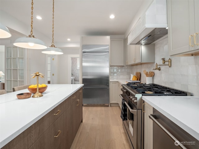 kitchen with high end appliances, custom range hood, white cabinets, decorative light fixtures, and backsplash