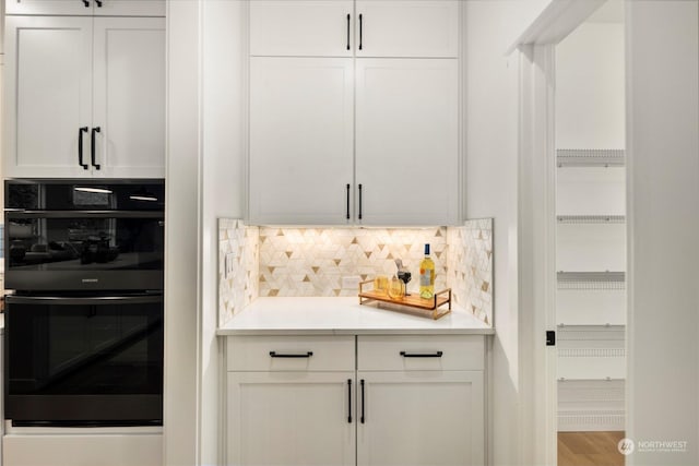 bar with black double oven, white cabinetry, wood-type flooring, and tasteful backsplash