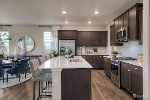 kitchen with appliances with stainless steel finishes, backsplash, sink, wood-type flooring, and an island with sink