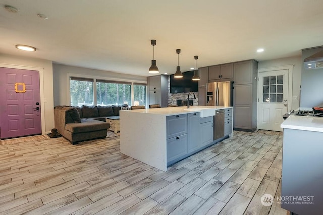 kitchen with gray cabinetry, stainless steel appliances, sink, hanging light fixtures, and an island with sink