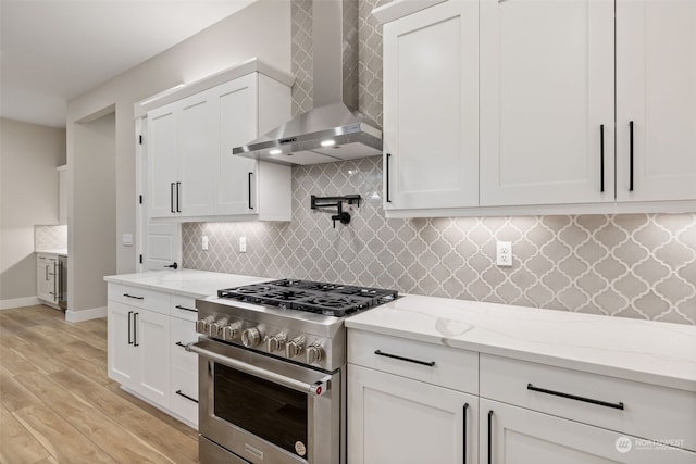 kitchen featuring white cabinets, stainless steel stove, wall chimney exhaust hood, light stone countertops, and light hardwood / wood-style floors