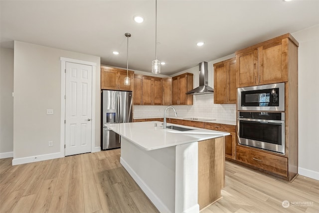 kitchen featuring wall chimney range hood, sink, appliances with stainless steel finishes, an island with sink, and decorative light fixtures