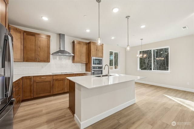 kitchen featuring sink, decorative light fixtures, fridge, an island with sink, and wall chimney range hood