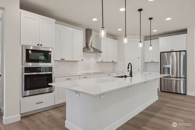 kitchen with a kitchen island with sink, white cabinets, wall chimney range hood, sink, and appliances with stainless steel finishes