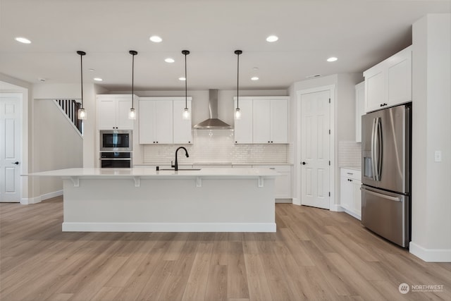 kitchen with white cabinetry, stainless steel appliances, wall chimney range hood, an island with sink, and decorative light fixtures