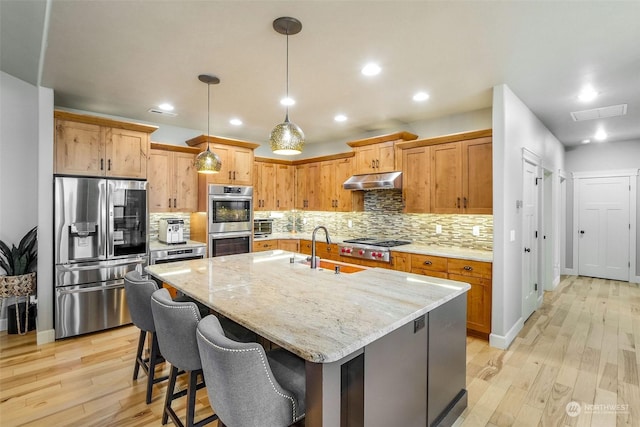 kitchen featuring a kitchen island with sink, appliances with stainless steel finishes, light stone counters, sink, and light hardwood / wood-style flooring