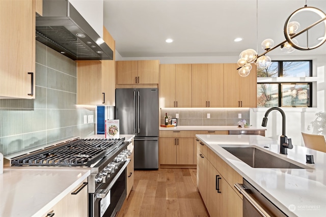 kitchen with wall chimney range hood, sink, hanging light fixtures, light brown cabinetry, and premium appliances