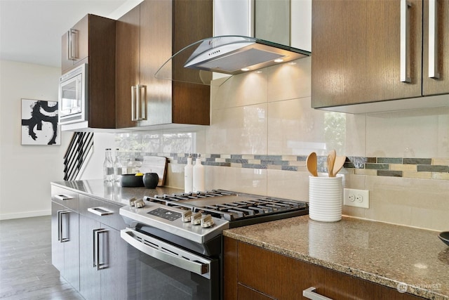 kitchen with backsplash, white microwave, stainless steel range with gas cooktop, wall chimney exhaust hood, and dark stone countertops