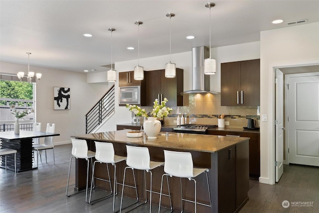 kitchen featuring decorative backsplash, wall chimney exhaust hood, dark stone countertops, stainless steel microwave, and an island with sink