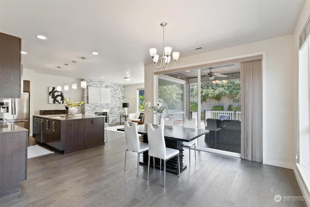 dining space with a stone fireplace, sink, dark wood-type flooring, and ceiling fan with notable chandelier