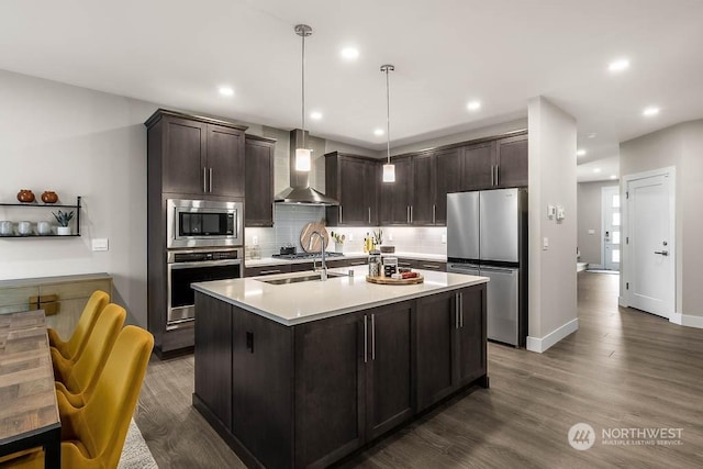 kitchen featuring dark brown cabinetry, stainless steel appliances, a kitchen island with sink, wall chimney range hood, and pendant lighting