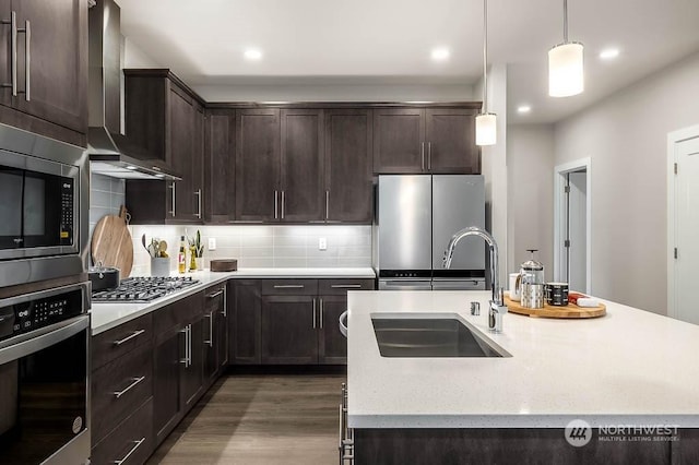 kitchen featuring appliances with stainless steel finishes, wall chimney exhaust hood, sink, hanging light fixtures, and an island with sink