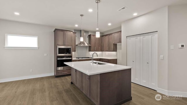 kitchen with appliances with stainless steel finishes, wall chimney exhaust hood, a center island with sink, decorative light fixtures, and dark hardwood / wood-style floors