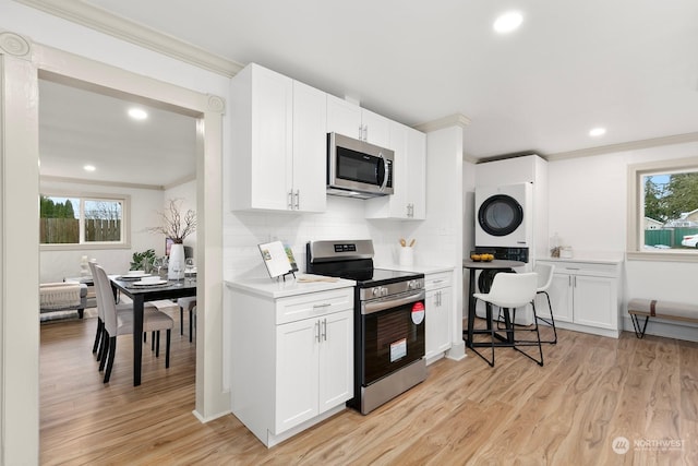 kitchen with appliances with stainless steel finishes, light wood-type flooring, tasteful backsplash, stacked washer and clothes dryer, and white cabinetry