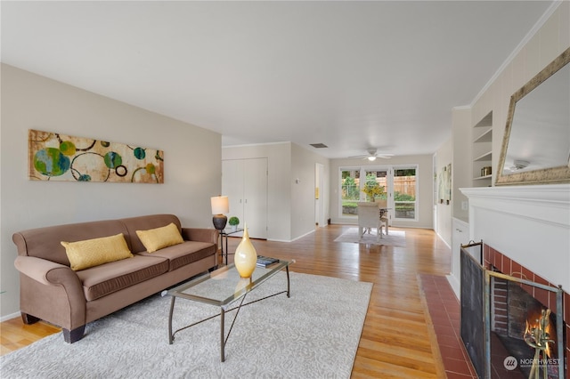 living room with a brick fireplace, ceiling fan, built in features, ornamental molding, and wood-type flooring