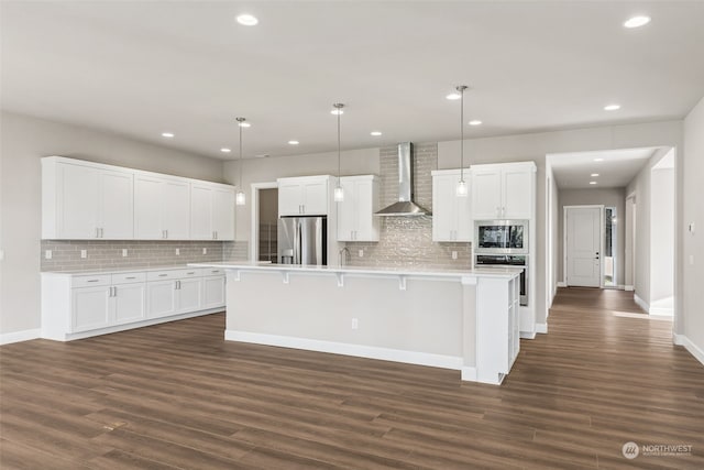 kitchen featuring appliances with stainless steel finishes, a kitchen island with sink, wall chimney range hood, white cabinetry, and hanging light fixtures