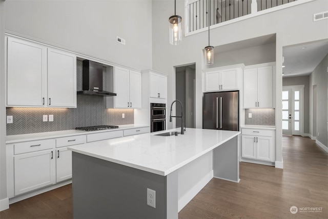 kitchen featuring white cabinetry, wall chimney exhaust hood, stainless steel appliances, decorative light fixtures, and a center island with sink