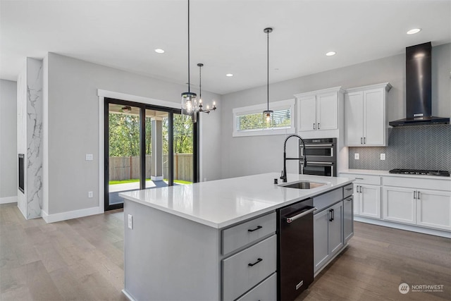 kitchen featuring white cabinets, sink, hanging light fixtures, wall chimney exhaust hood, and an island with sink