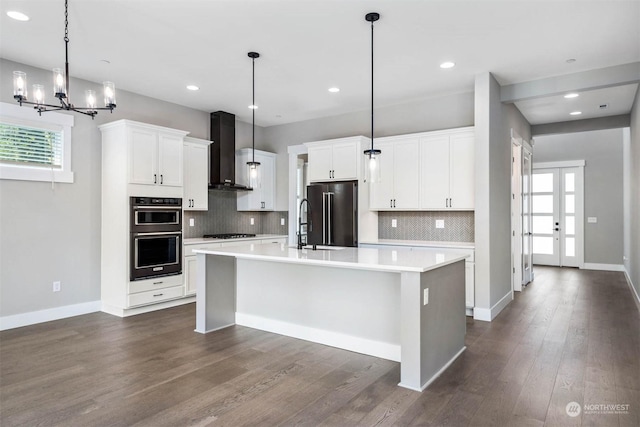 kitchen featuring hanging light fixtures, stainless steel appliances, wall chimney range hood, an island with sink, and white cabinets