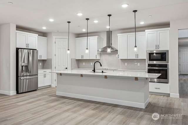 kitchen with appliances with stainless steel finishes, a kitchen island with sink, decorative light fixtures, wall chimney range hood, and white cabinets