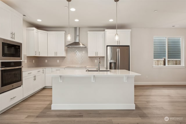 kitchen with white cabinets, decorative light fixtures, wall chimney range hood, stainless steel appliances, and a center island with sink