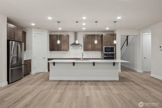 kitchen featuring wall chimney exhaust hood, stainless steel appliances, pendant lighting, and an island with sink