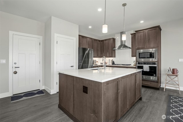 kitchen featuring a center island with sink, wall chimney range hood, hanging light fixtures, dark brown cabinets, and stainless steel appliances