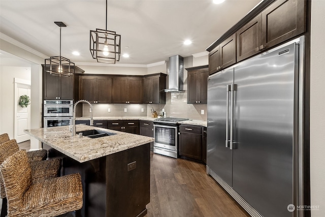 kitchen with a kitchen island with sink, sink, wall chimney exhaust hood, appliances with stainless steel finishes, and decorative light fixtures