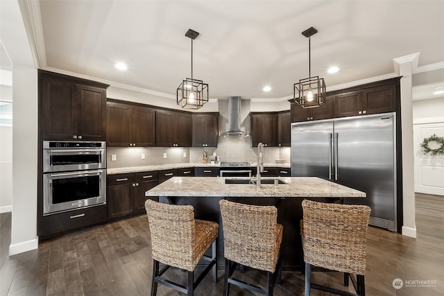 kitchen with sink, wall chimney exhaust hood, hanging light fixtures, stainless steel appliances, and an island with sink