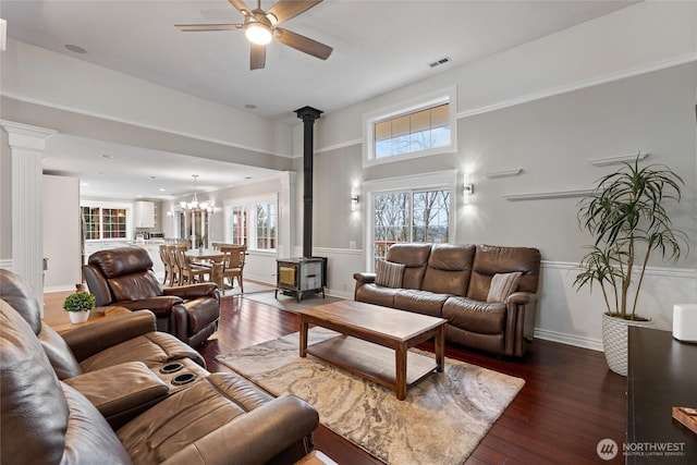 living area featuring ceiling fan with notable chandelier, dark wood-type flooring, visible vents, wainscoting, and a wood stove