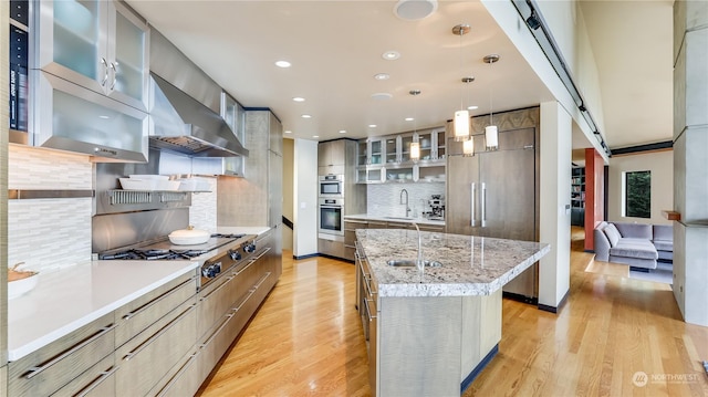 kitchen featuring a center island, hanging light fixtures, glass insert cabinets, a sink, and wall chimney range hood