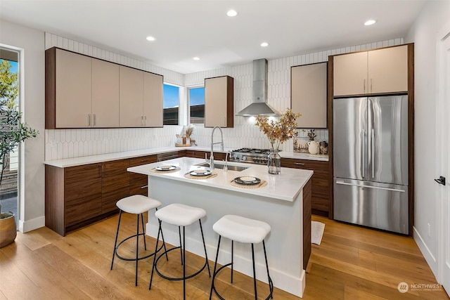 kitchen with a kitchen island with sink, wall chimney range hood, sink, a kitchen bar, and stainless steel refrigerator