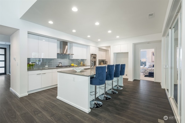 kitchen with dark wood-type flooring, a center island with sink, wall chimney exhaust hood, white cabinetry, and stainless steel appliances