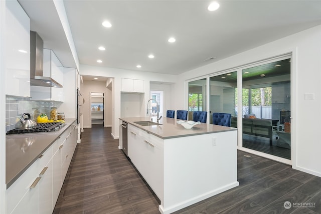 kitchen featuring sink, stainless steel appliances, wall chimney range hood, an island with sink, and white cabinets