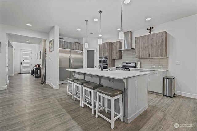 kitchen with pendant lighting, a kitchen island with sink, wall chimney exhaust hood, decorative backsplash, and appliances with stainless steel finishes