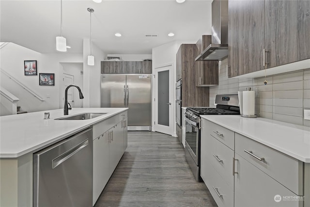 kitchen with appliances with stainless steel finishes, wall chimney exhaust hood, sink, hanging light fixtures, and an island with sink